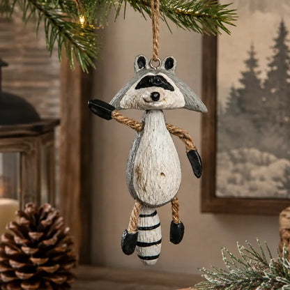 A hanging raccoon Christmas ornament made to look like a distressed wood carving, suspended from a pine branch in a rustic indoor setting with a pinecone and framed picture in the background.